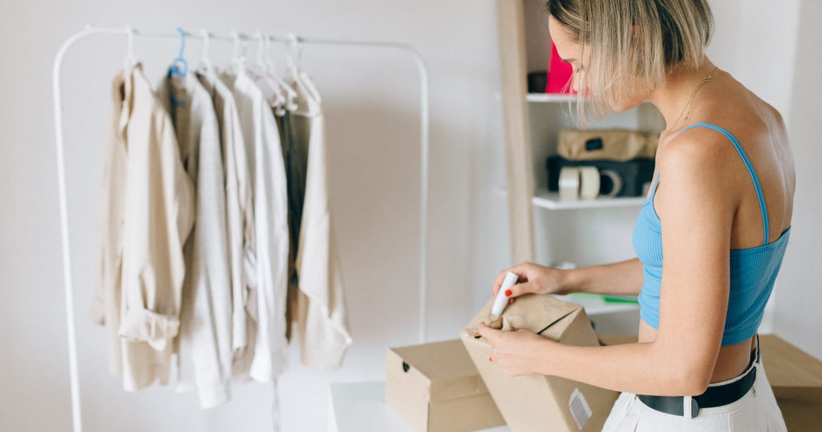 Persona cerrando una caja de envío junto a un perchero de ropa, con la política de cambios y devoluciones de su tienda online en mente